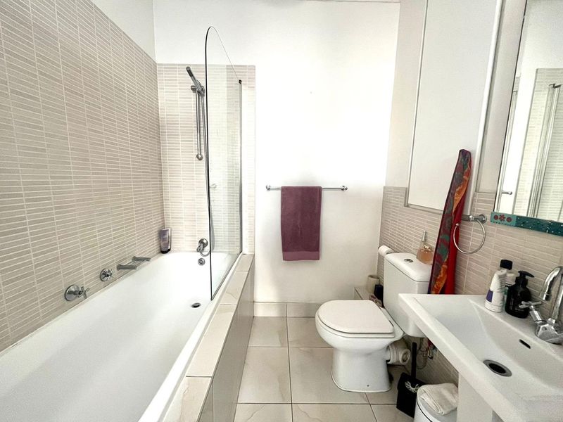 A clean, functional bathroom featuring a built-in bathtub with a glass screen, pedestal washbasin, and neutral mosaic wall tiling.