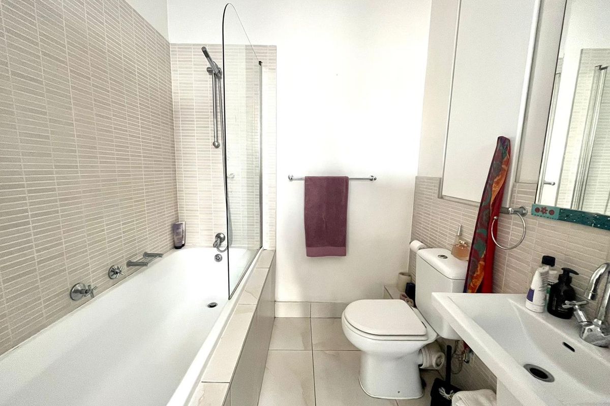 A clean, functional bathroom featuring a built-in bathtub with a glass screen, pedestal washbasin, and neutral mosaic wall tiling.