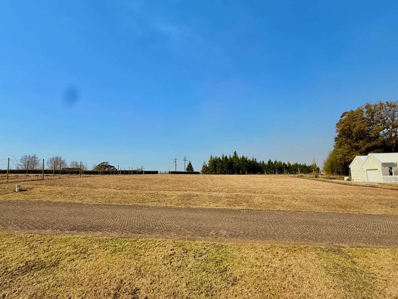 A wide, level residential vacant land plot featuring a paved driveway, perimeter wire fencing, and established trees in a quiet setting.