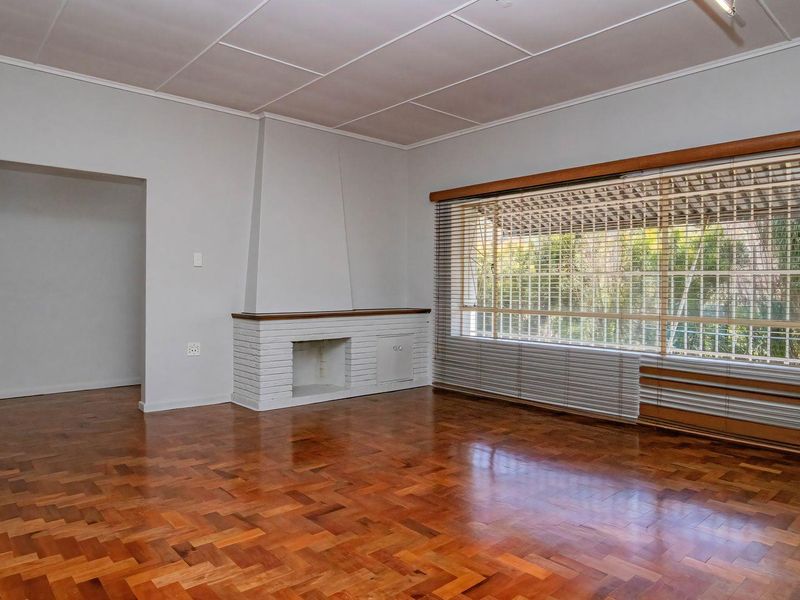 Spacious living room featuring classic parquet flooring, a built-in brick fireplace, and a large window with venetian blinds overlooking the garden.