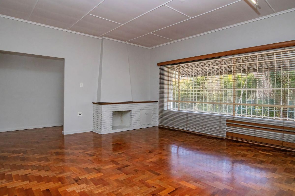 Spacious living room featuring classic parquet flooring, a built-in brick fireplace, and a large window with venetian blinds overlooking the garden.