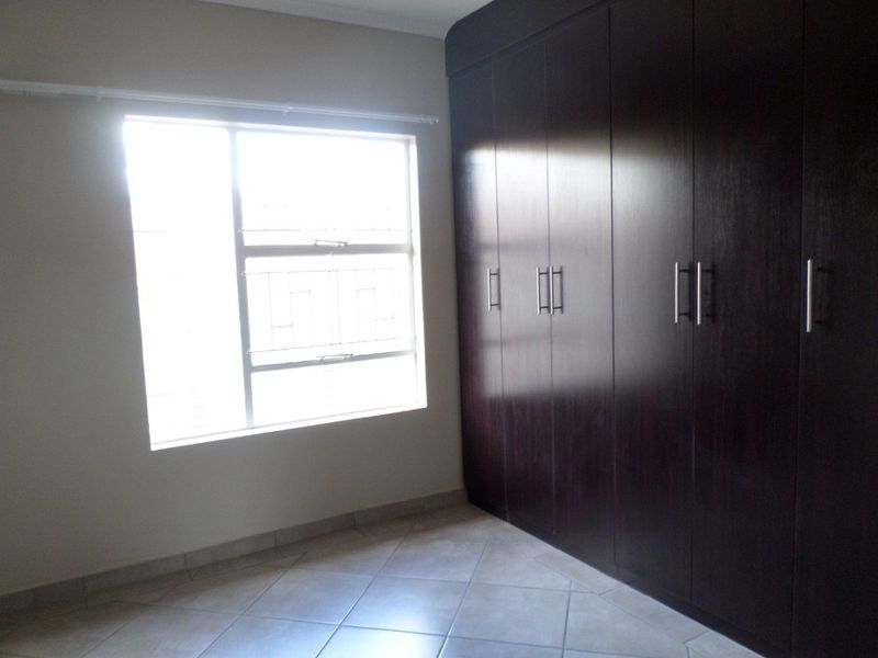 A bright, empty bedroom featuring dark wood built-in cupboards, tiled flooring, and a window with security bars.