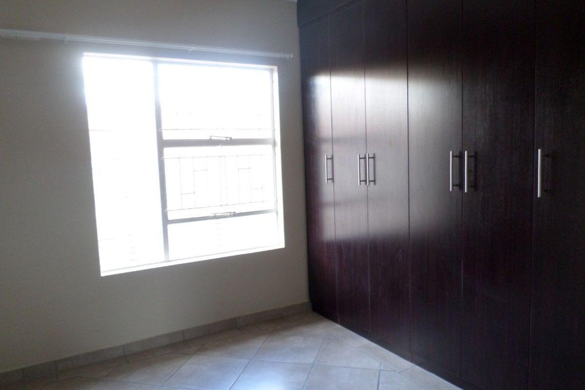 A bright, empty bedroom featuring dark wood built-in cupboards, tiled flooring, and a window with security bars.