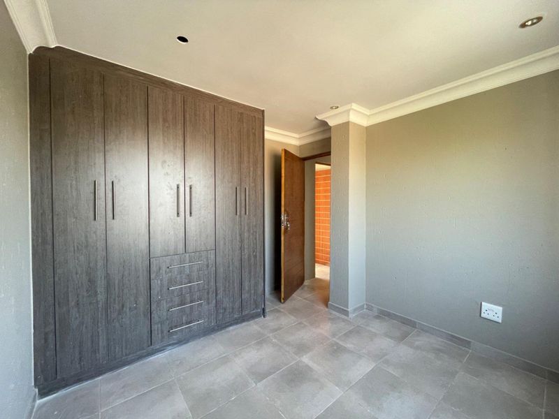 A clean, empty bedroom featuring modern dark wood built-in cupboards, neutral grey walls, tiled flooring, and recessed ceiling lighting.