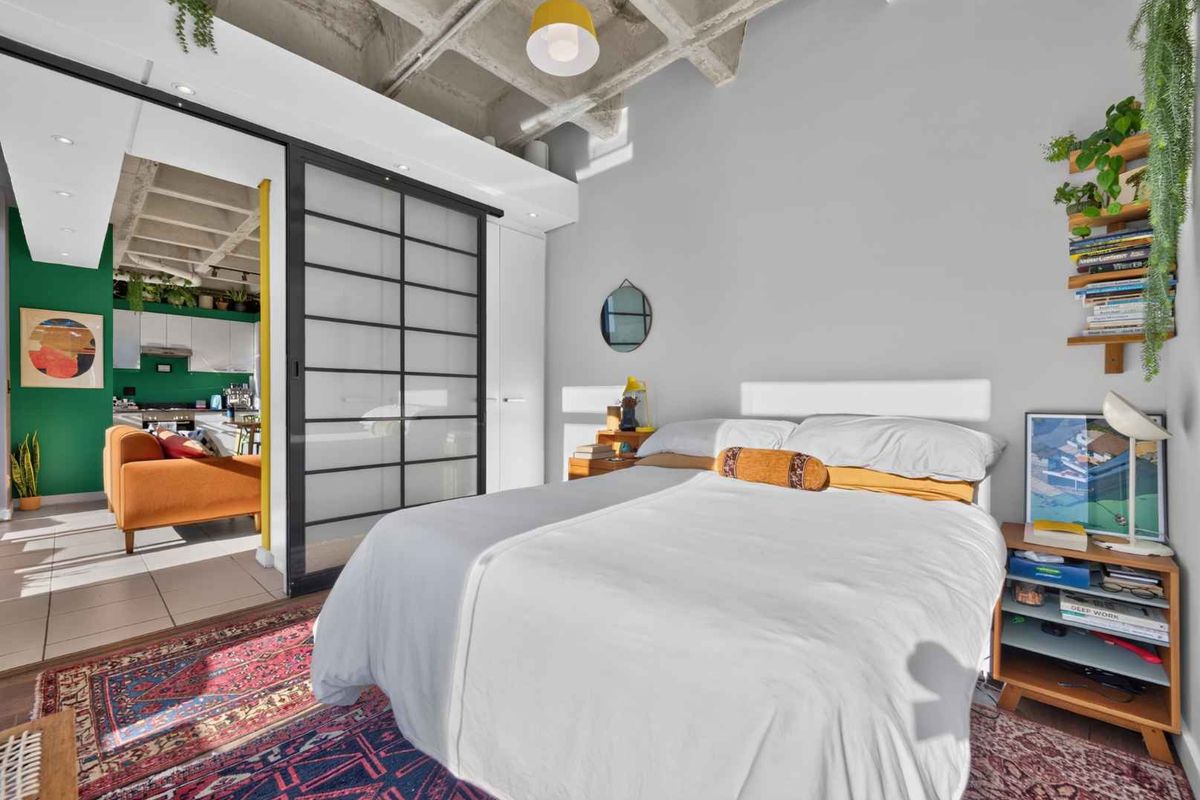 Modern bedroom featuring an exposed concrete waffle ceiling, sliding room divider, and built-in storage, with an adjacent view into the kitchen area.
