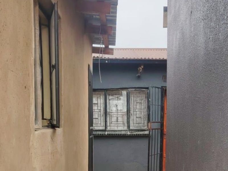 A narrow exterior side passage between two structures featuring basic masonry walls, security gates, and exposed roof rafters.