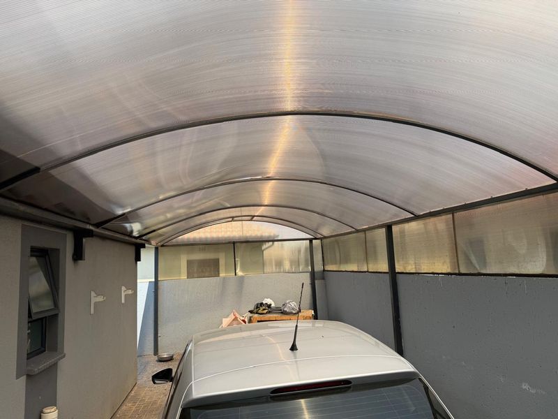 A covered carport area featuring a durable arched polycarbonate roof and metal support structure, providing sheltered parking space.