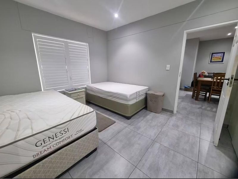 A bedroom featuring neutral grey walls, large format floor tiles, and shutter blinds, with an adjacent view into the dining area.