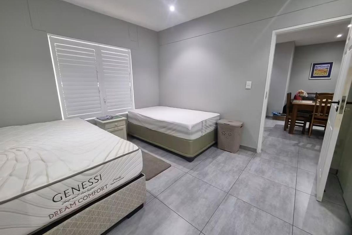 A bedroom featuring neutral grey walls, large format floor tiles, and shutter blinds, with an adjacent view into the dining area.