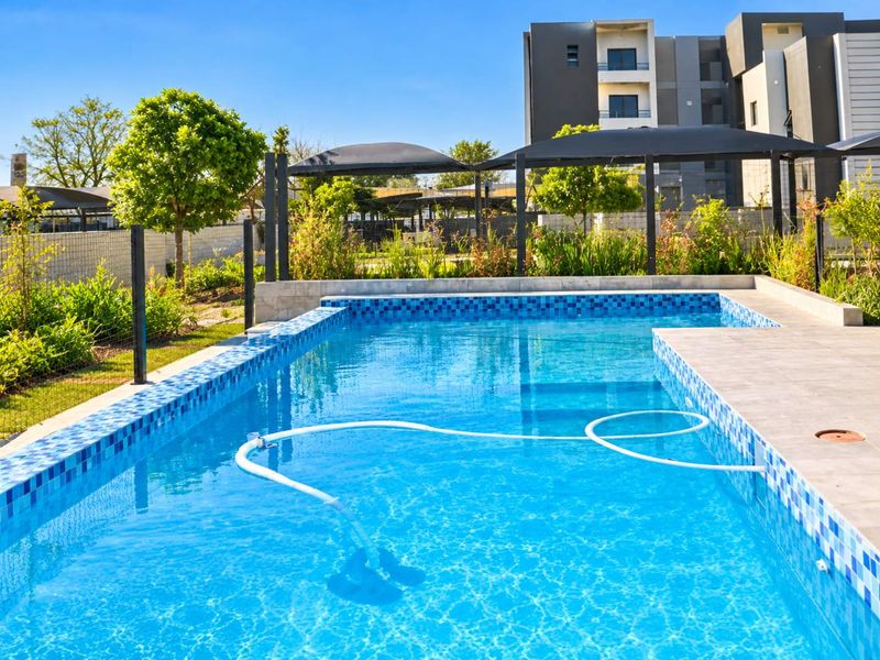 A well-maintained communal swimming pool area featuring tiled surrounds, shade sails, and landscaped garden borders within an apartment complex.