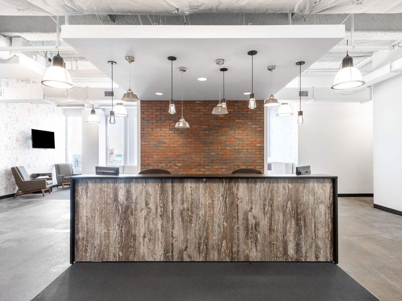 Modern office reception area featuring a wood-finish desk, exposed industrial ceiling, brick accent wall, and stylish pendant lighting.