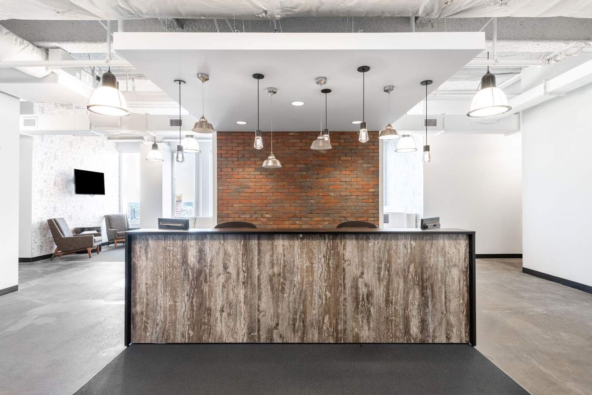 Modern office reception area featuring a wood-finish desk, exposed industrial ceiling, brick accent wall, and stylish pendant lighting.