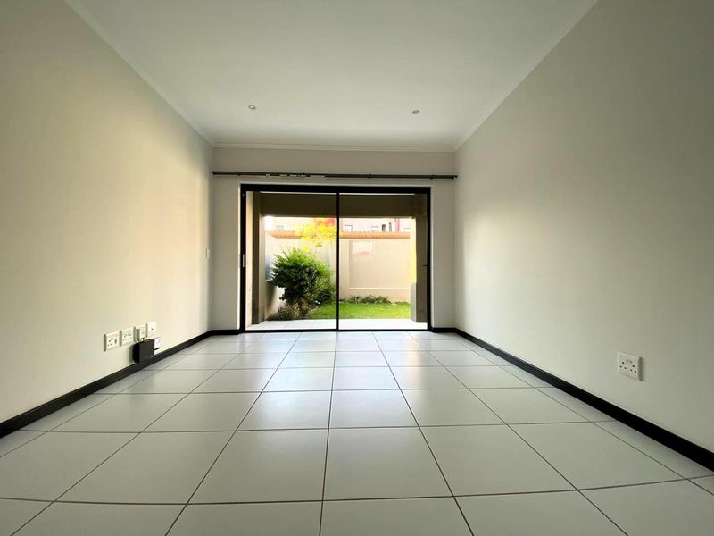 A bright, empty living room featuring neutral tiled flooring, recessed lighting, and a large sliding glass door providing access to a private rear garden.