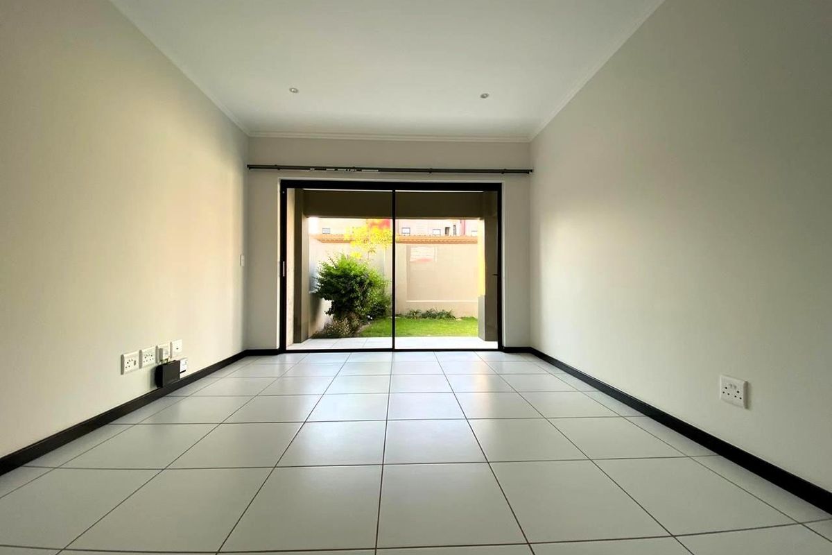 A bright, empty living room featuring neutral tiled flooring, recessed lighting, and a large sliding glass door providing access to a private rear garden.