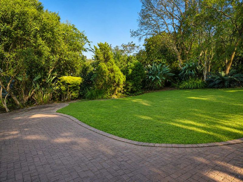 A well-maintained garden featuring a lush green lawn, mature trees, and a curved brick-edged paved walkway.