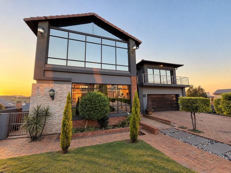 Modern double-story residence featuring a striking glass facade, stone cladding, brick-paved driveway, and an upper-level balcony with contemporary architectural design.