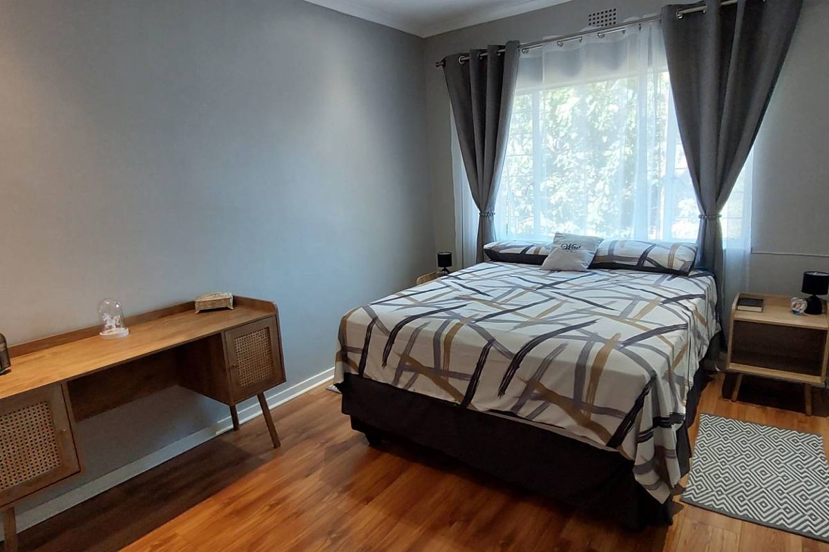 A well-maintained bedroom featuring warm wood-look flooring, neutral grey walls, and a large window with integrated curtain tracks.