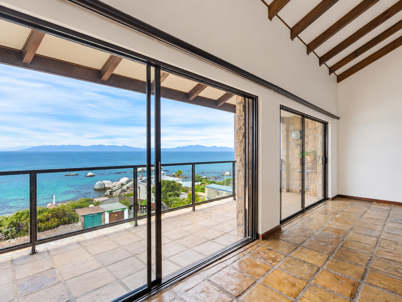 Spacious covered balcony featuring exposed timber rafters, stone flooring, and expansive sliding glass doors offering panoramic ocean and coastal views.