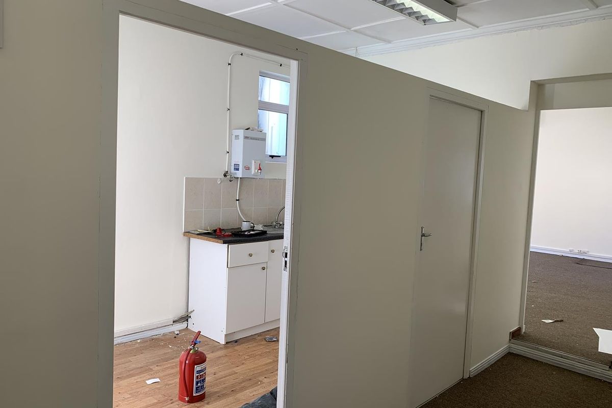 A functional office kitchenette area featuring built-in cabinetry, a sink, and a wall-mounted water heater, adjacent to a carpeted office workspace.