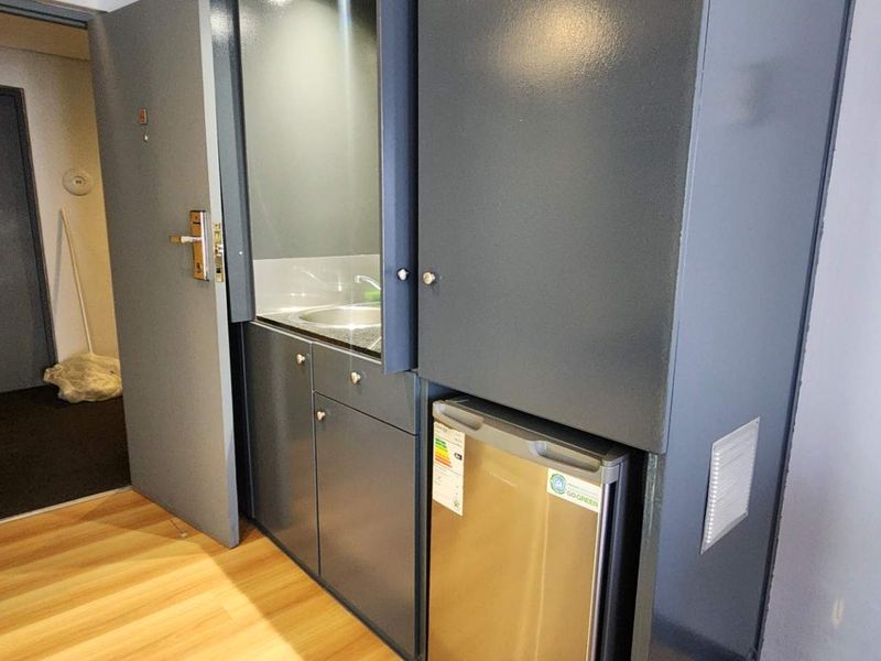 A compact kitchenette featuring built-in cabinetry, a stainless steel sink, and an integrated under-counter refrigerator, located in a hallway area.