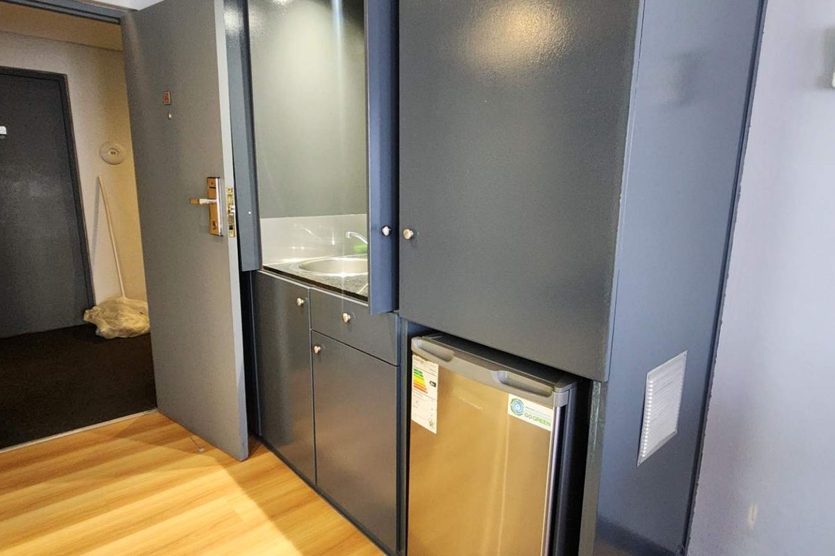 A compact kitchenette featuring built-in cabinetry, a stainless steel sink, and an integrated under-counter refrigerator, located in a hallway area.