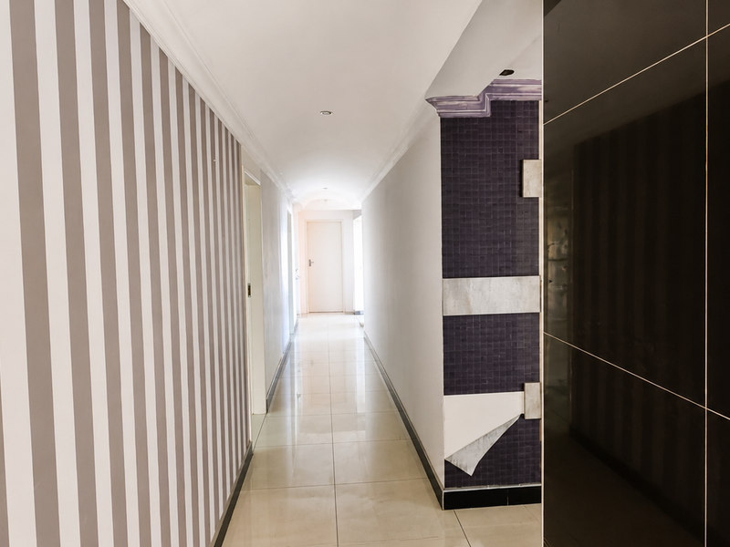 A long, tiled hallway featuring decorative striped wallpaper and a contrasting tiled pillar, leading to a bright doorway at the end.
