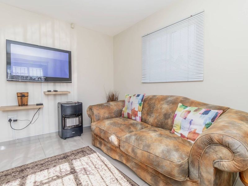 A compact living room featuring tiled flooring, wall-mounted shelving, and venetian blinds, providing a functional and neutral space.