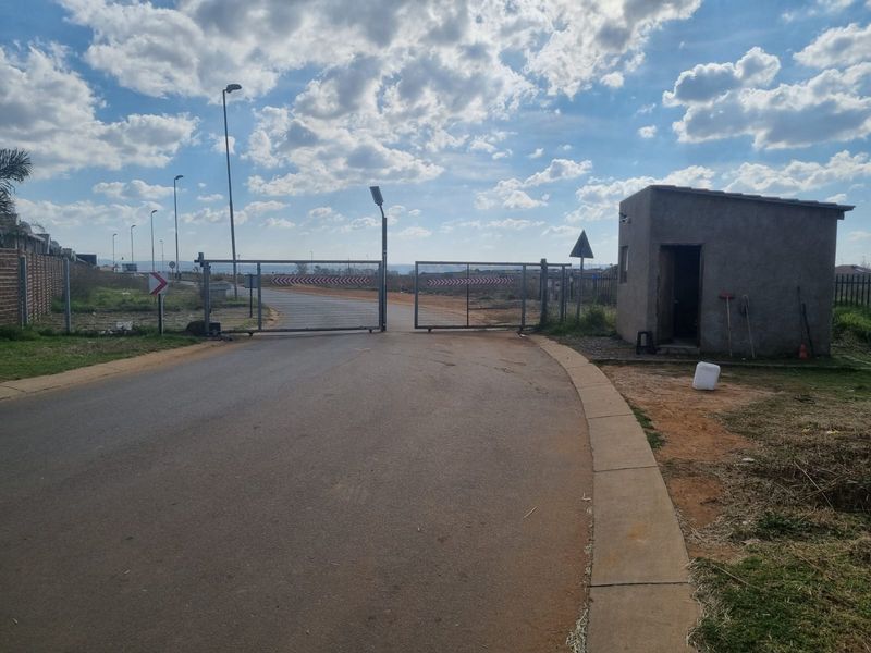 Entrance to the property featuring a security gate, guard house, and asphalt access road within a developing residential area.