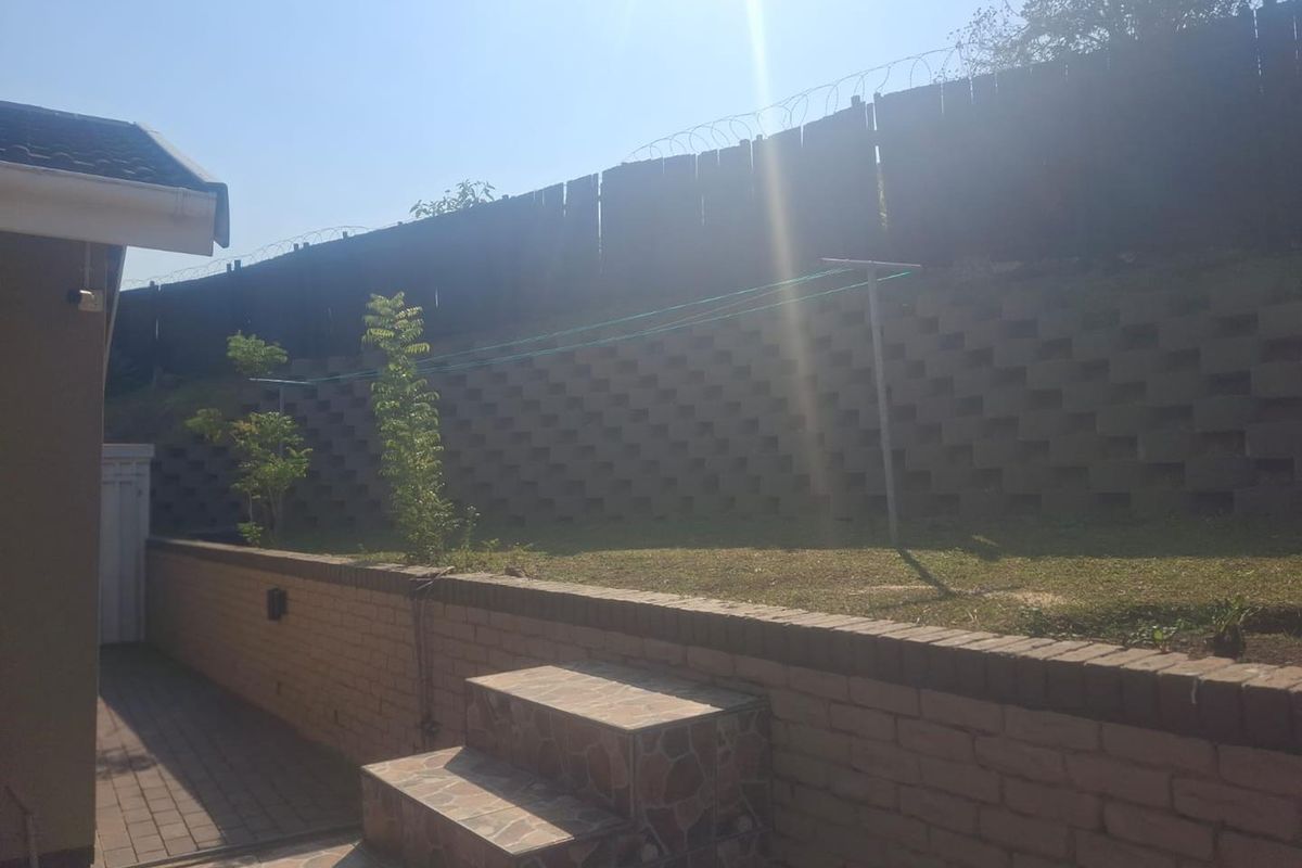 Rear exterior view featuring a tiered garden with a retaining wall, brick boundary wall with security fencing, and a paved walkway with tiled steps.