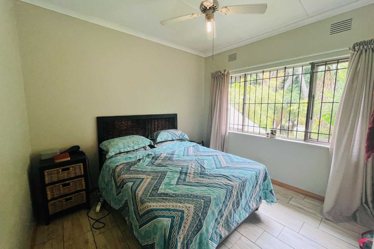 A bedroom featuring tiled flooring, a ceiling fan, and a large window with security bars overlooking the garden.
