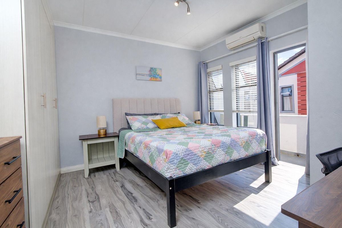 A well-lit bedroom featuring built-in cupboards, laminate flooring, a wall-mounted air conditioning unit, and direct access to a private balcony.
