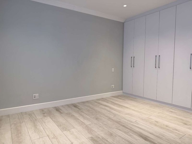 A bright, modern bedroom featuring light-toned laminate flooring, neutral grey walls, and extensive built-in floor-to-ceiling cupboards for ample storage.