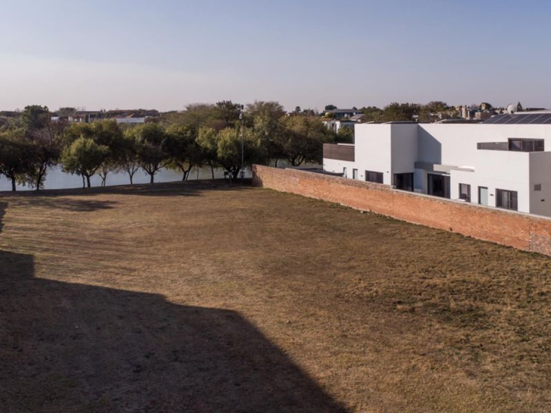 A spacious vacant residential plot featuring a brick boundary wall, adjacent to a modern residential building with a view of nearby trees and water.