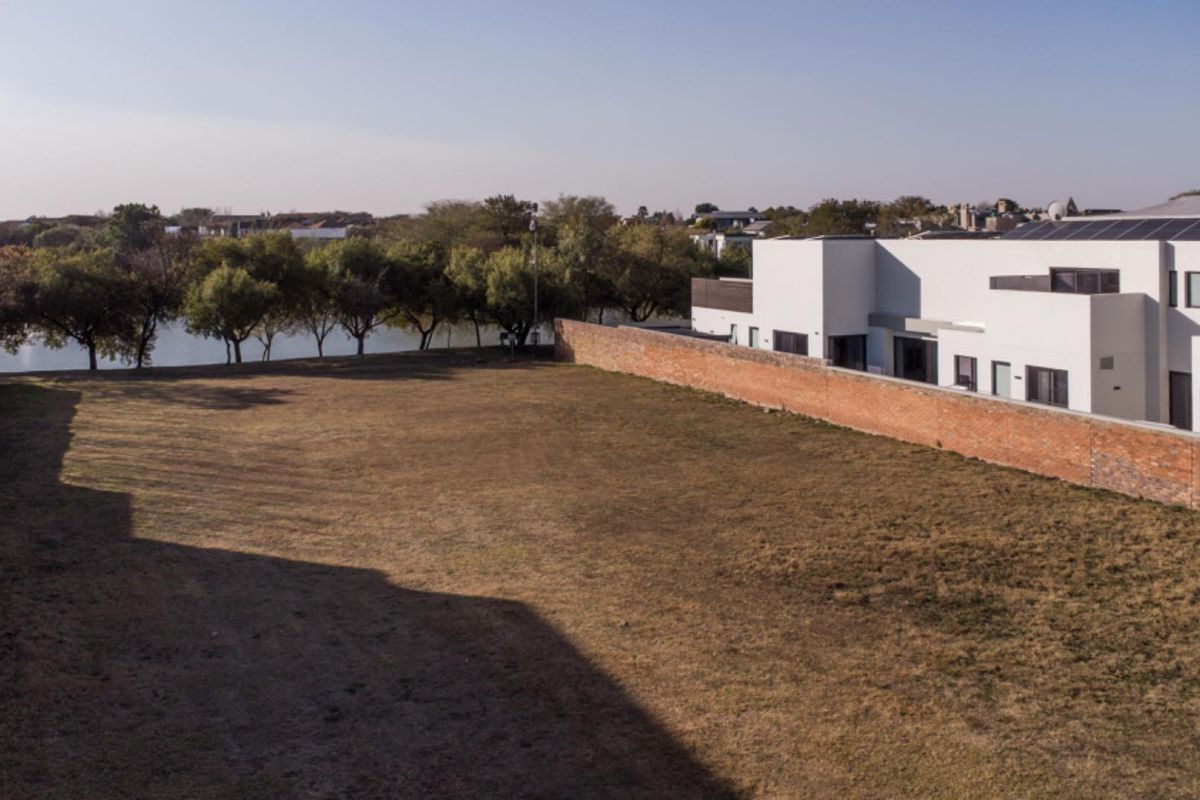 A spacious vacant residential plot featuring a brick boundary wall, adjacent to a modern residential building with a view of nearby trees and water.