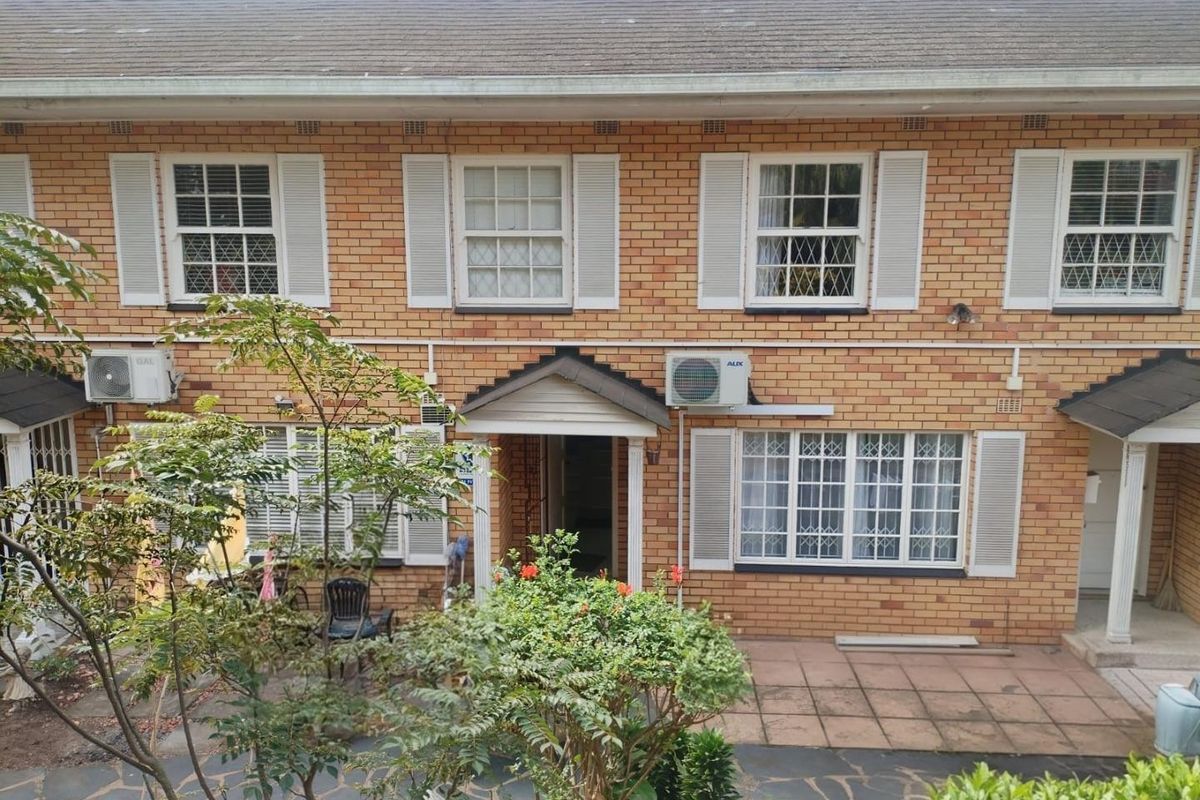Rear view of a brick townhouse featuring a paved patio area, multiple sash windows with security shutters, and wall-mounted air conditioning units.