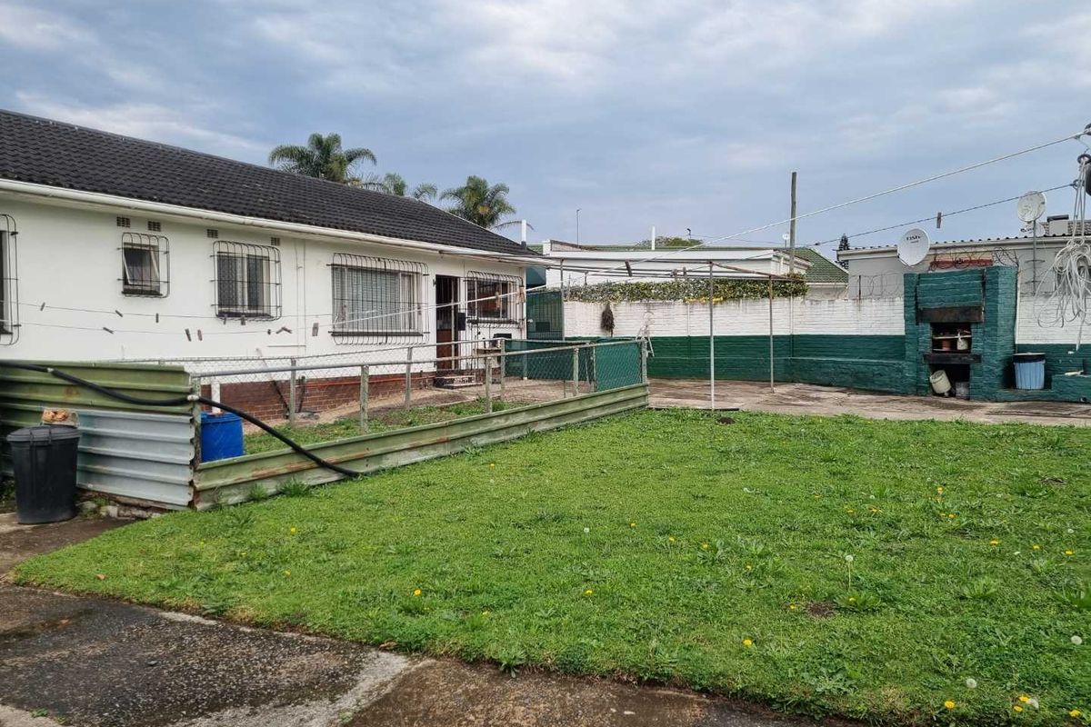 Rear exterior view of the property featuring a lawn, concrete patio area, built-in braai, and perimeter walling with security features.