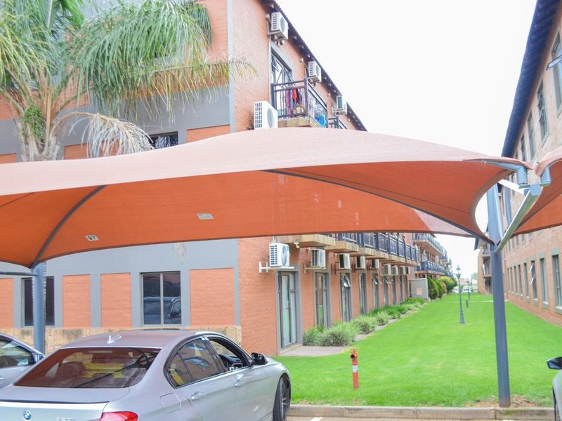 Exterior view of an apartment complex featuring brick facades, multiple balconies with air conditioning units, and a covered parking area with shade sails.