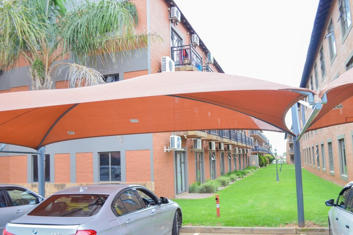 Exterior view of an apartment complex featuring brick facades, multiple balconies with air conditioning units, and a covered parking area with shade sails.