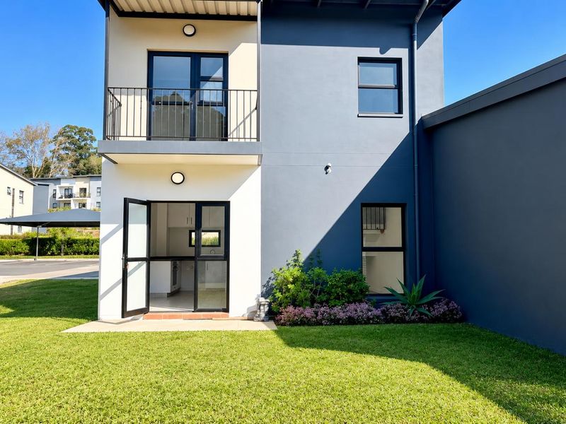 Rear view of a modern two-story townhouse featuring a private garden, a balcony with metal railing, and large glass sliding doors.