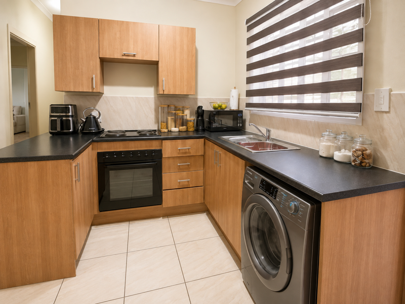 A functional kitchen featuring light wood-look cabinetry, dark laminate countertops, tiled backsplash, and integrated appliances including an oven and hob.
