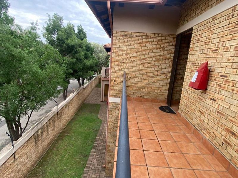 A covered balcony featuring tiled flooring, brick walls, and a metal safety railing, overlooking the ground level garden area.