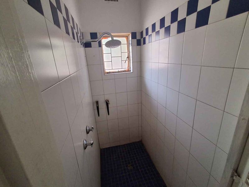 A compact shower stall featuring white wall tiles with a blue checkered border, dark floor tiles, and a small window with security bars.