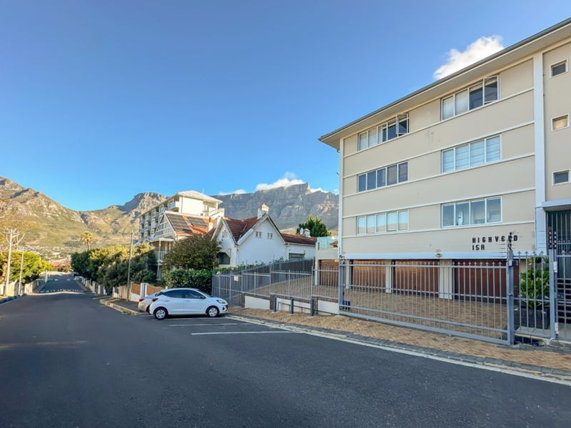Front exterior view of a multi-level apartment building featuring a paved driveway, secure metal gate, and mountain views in the background.
