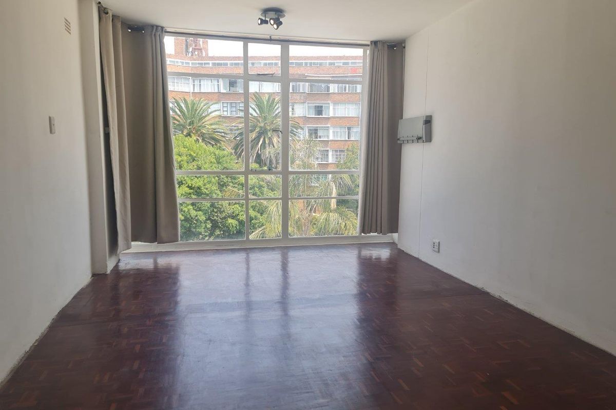 A bright, empty living room featuring polished parquet flooring and a large floor-to-ceiling window with views of the surrounding neighborhood.