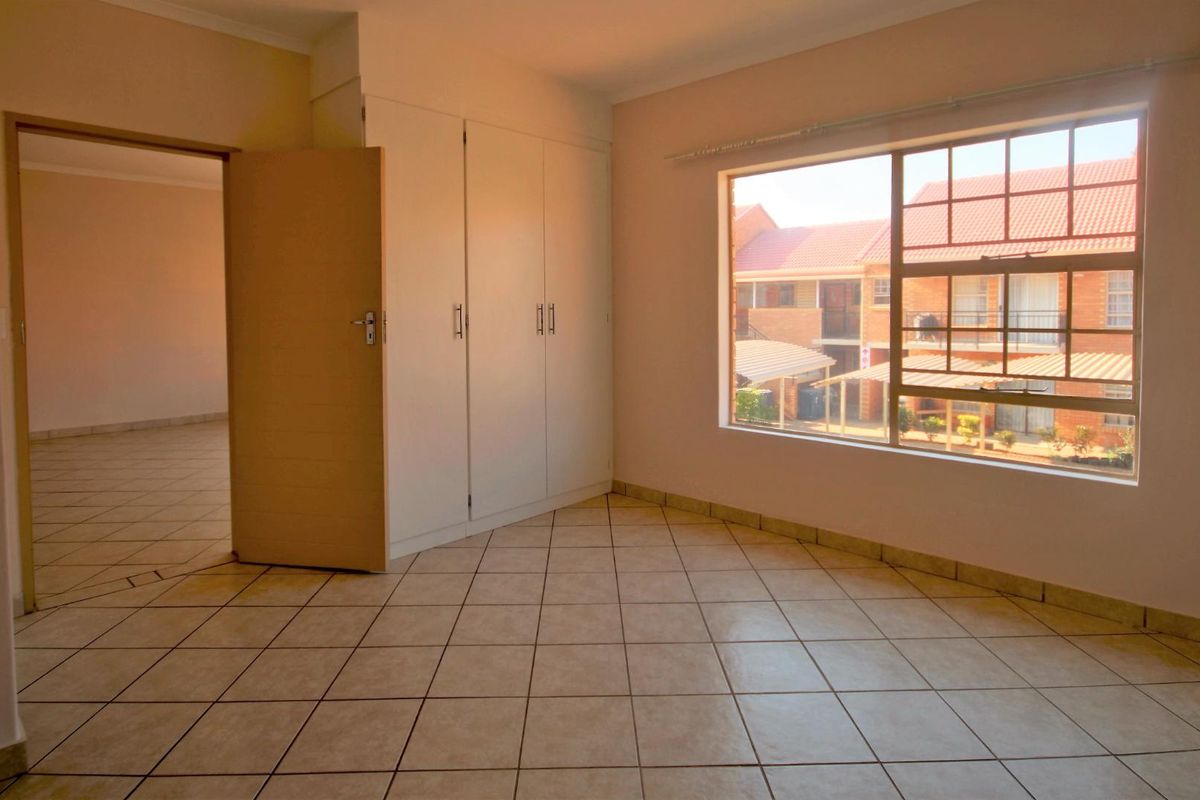 A bright, empty bedroom featuring neutral tiled flooring, built-in cupboards, and a large window overlooking the apartment complex.