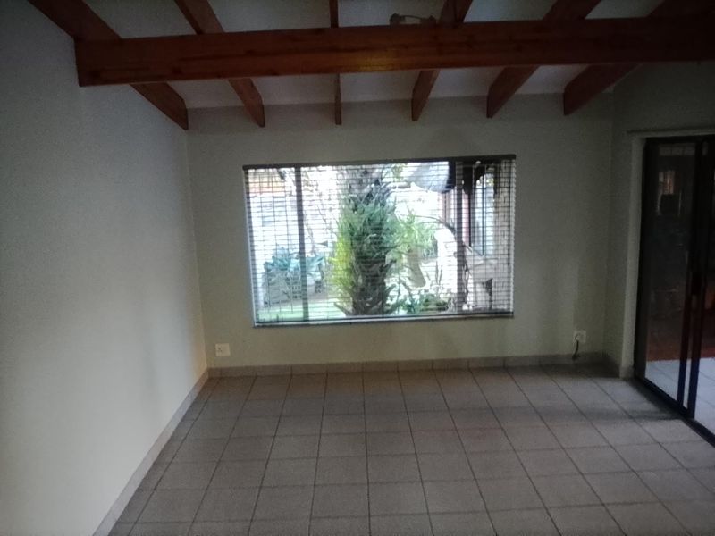 An empty living room featuring exposed timber ceiling beams, tiled flooring, and a large window overlooking the garden.