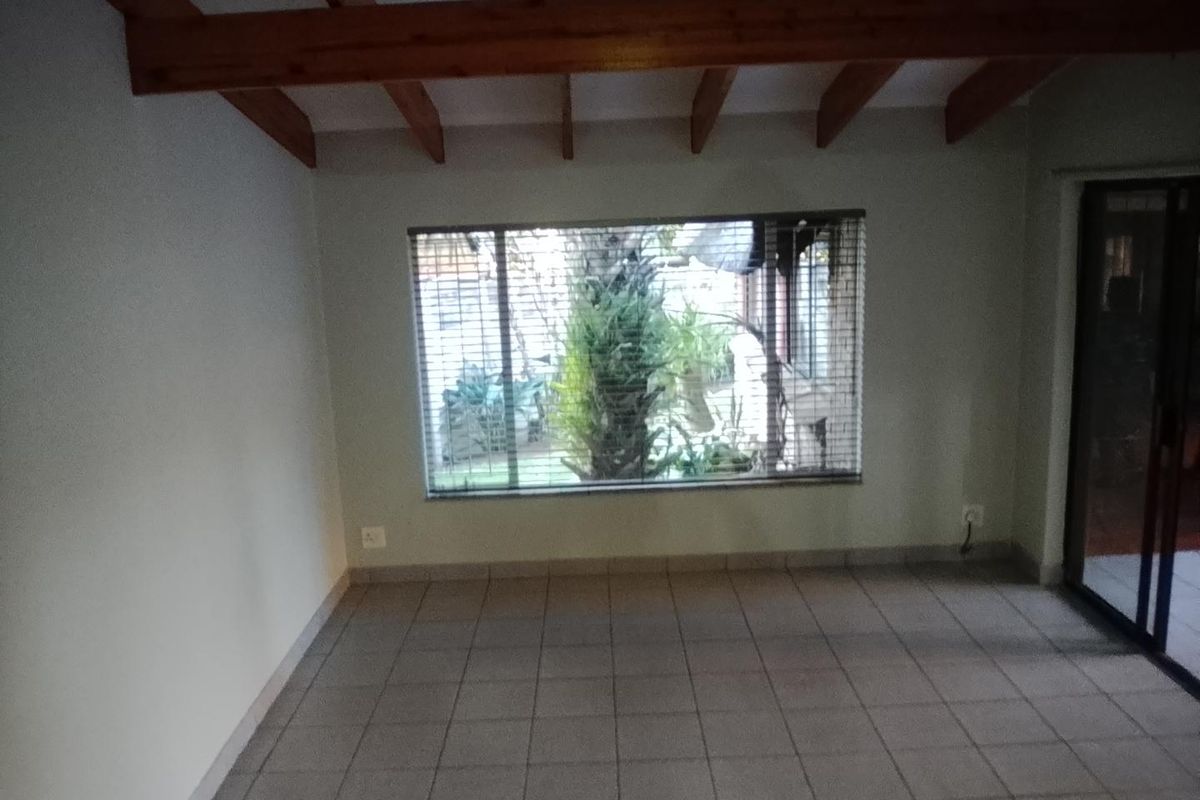 An empty living room featuring exposed timber ceiling beams, tiled flooring, and a large window overlooking the garden.