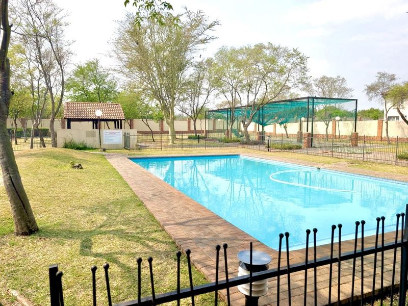 Outdoor pool area featuring a swimming pool with brick coping, metal fencing, a small thatched gazebo, and a shaded sports enclosure in the garden.