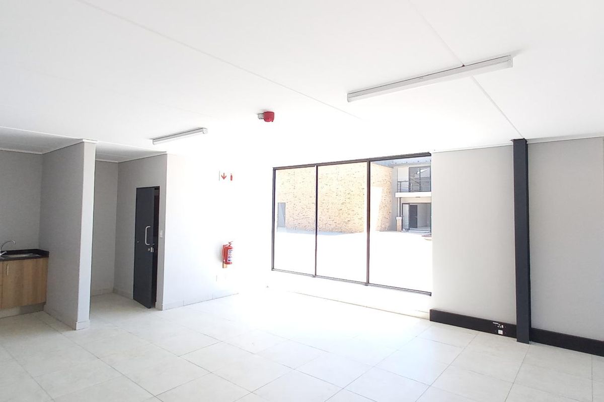 A bright, open-plan industrial office space featuring tiled flooring, a small kitchenette area, and large windows providing ample natural light.