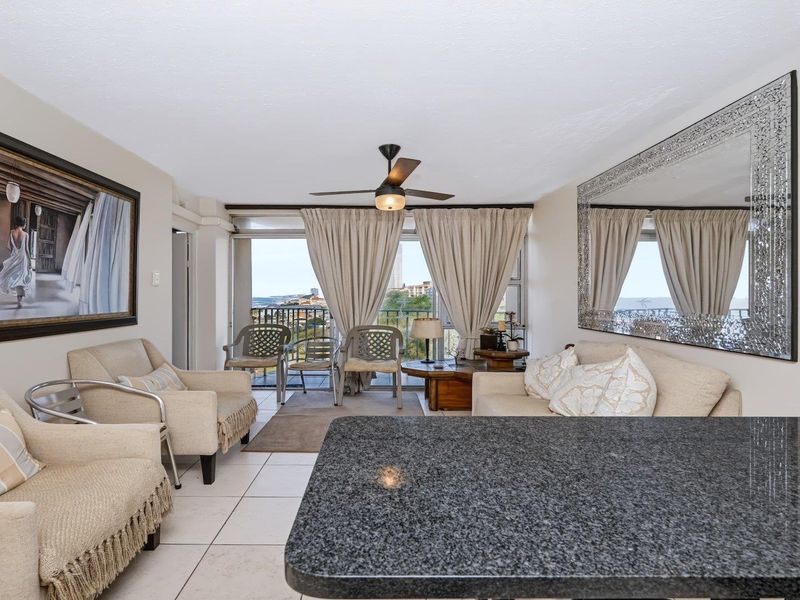 Open-plan living area featuring tiled flooring, a ceiling fan, and a granite breakfast bar, with direct access to a balcony offering scenic views.